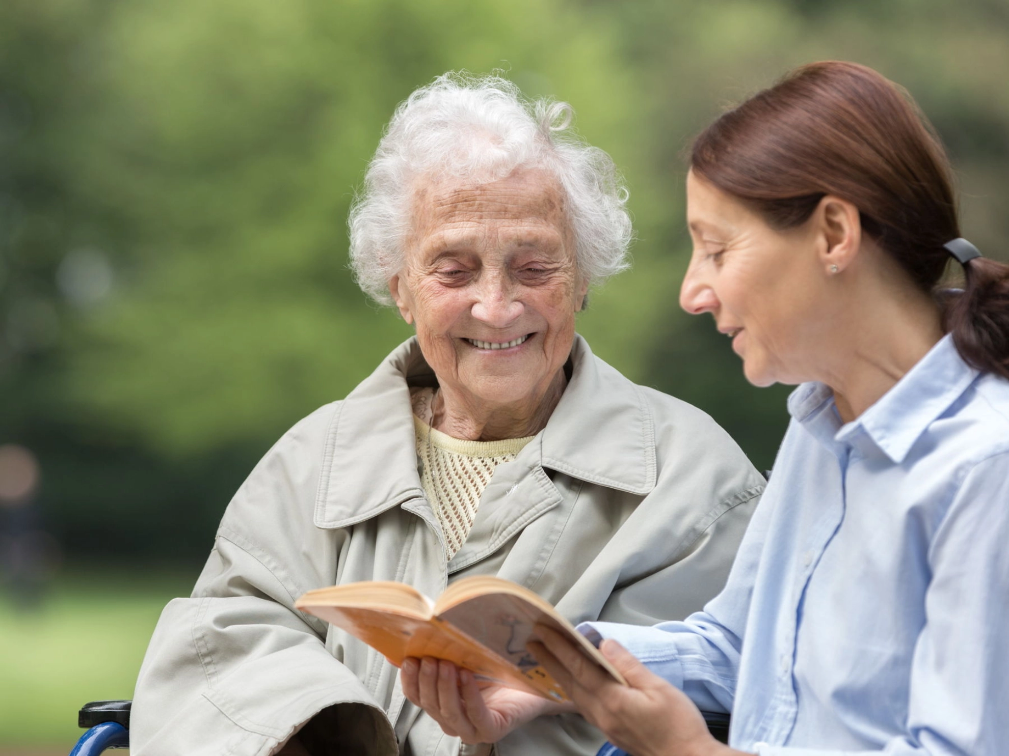 Eine Seniorin im Rollstuhl und ihre Tochter schauen in einem Park gemeinsam in ein Buch.